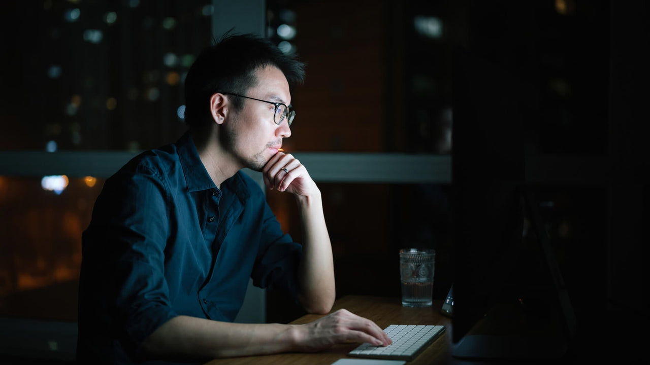 Man sitting at a desk at night looking at a computer monitor.