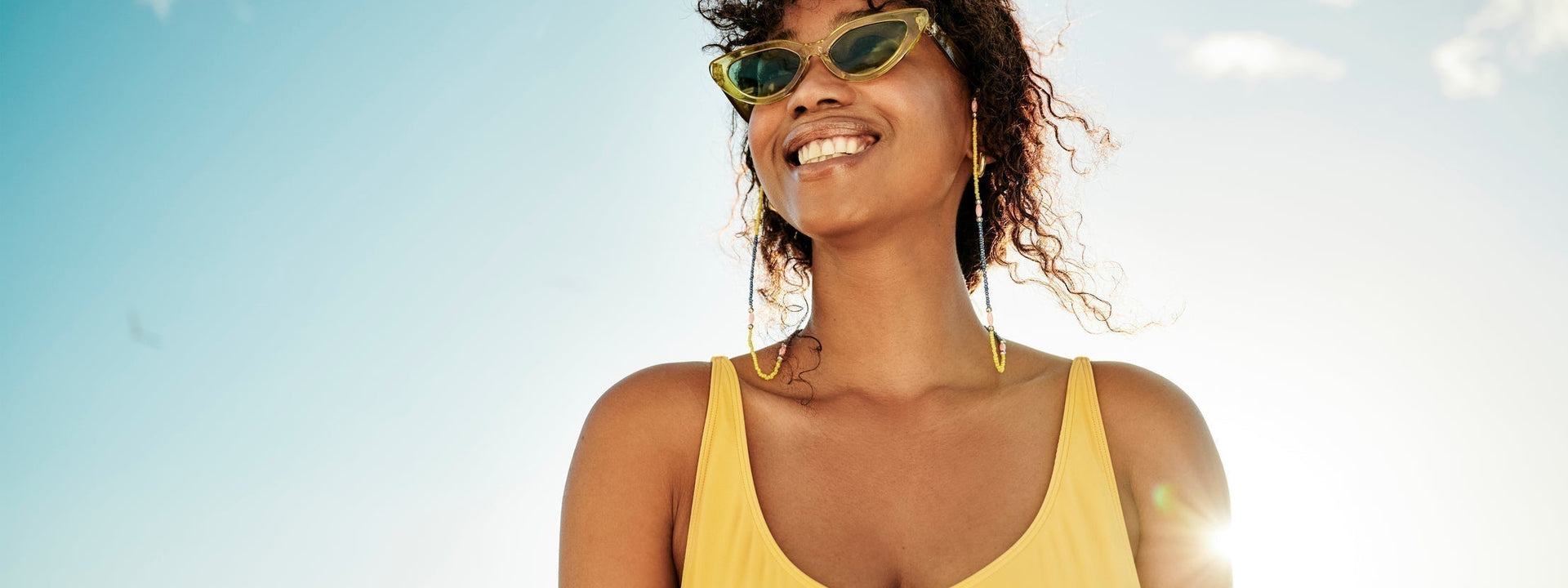 A woman in a yellow tank top smiling against a sunny blue sky background.