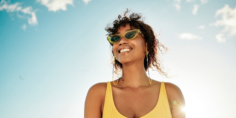 A woman in a yellow tank top smiling against a sunny blue sky background.
